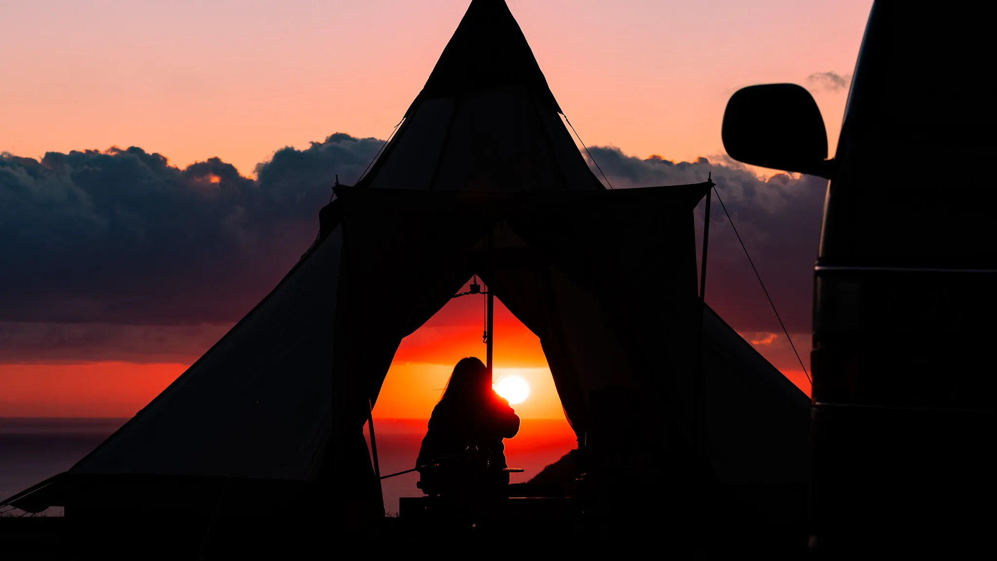 夕日の中、テントの入り口で景色を眺めるキャンパーのシルエット